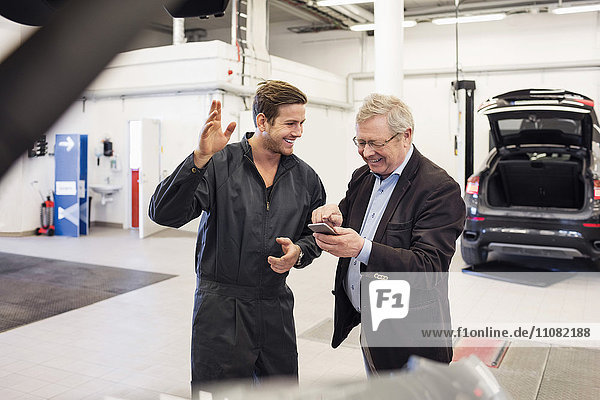 Fröhlicher Mechaniker mit Senioren mit mobiler App in der Autowerkstatt