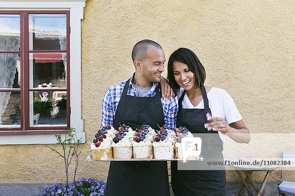 Smiling bakers holding tray of pastries while standing against wall outside cafe