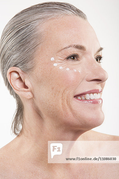 Nahaufnahme einer fröhlichen reifen Frau mit Punkten aus Feuchtigkeitscreme auf dem Gesicht vor weißem Hintergrund