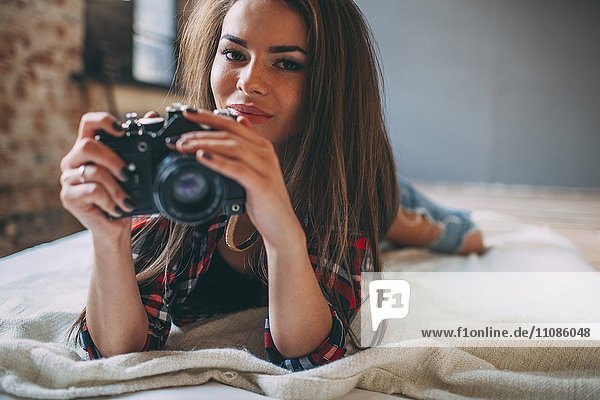 Portrait of young woman holding SLR camera while lying on bed at home