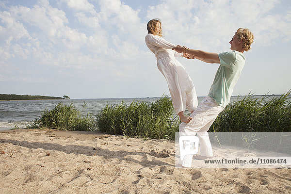 Friend balancing on man's knee at beach against sky