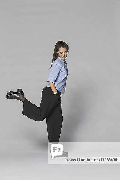 Durchgehende Frau auf einem Bein stehend mit Händen in Taschen vor grauem Hintergrund