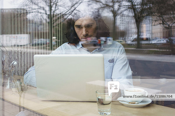Man using laptop and having coffee seen through glass window