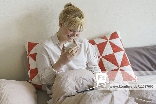 Lächelnde Frau beim Kaffeetrinken mit dem digitalen Tablett im Bett zu Hause