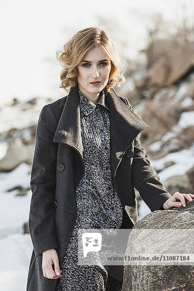 Portrait of beautiful young woman wearing long coat while standing by rock