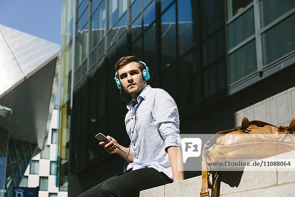 Irland  Dublin  junger Geschäftsmann auf einer Wand sitzend Musik hören mit Kopfhörern