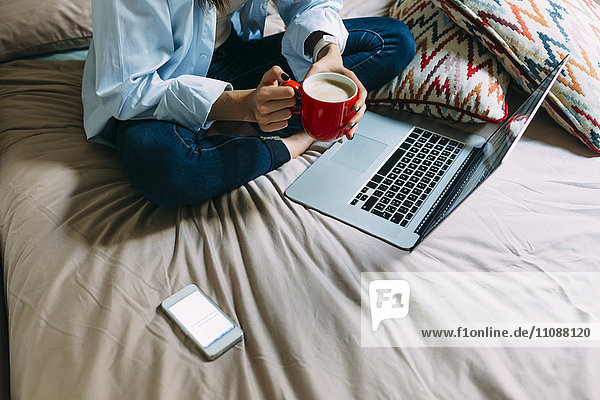 Junge Frau sitzt auf Bett  Kaffeetasse  Laptop und Smartphone