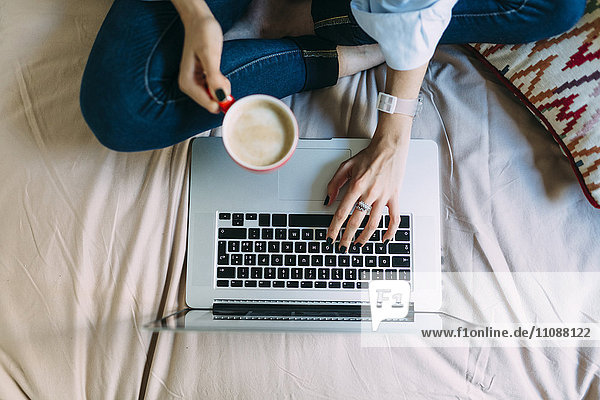Junge Frau sitzt auf Bett Kaffeetasse Laptop und Smartphone