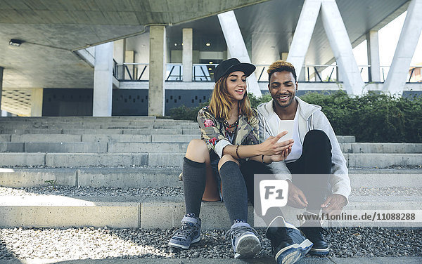 Lächelndes junges Paar sitzt auf einer Treppe und schaut auf das Smartphone.
