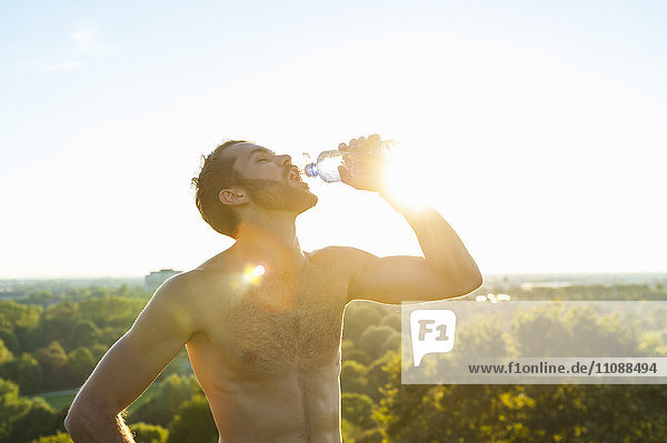 Barechested Athlet  der bei Sonnenuntergang aus der Flasche trinkt.