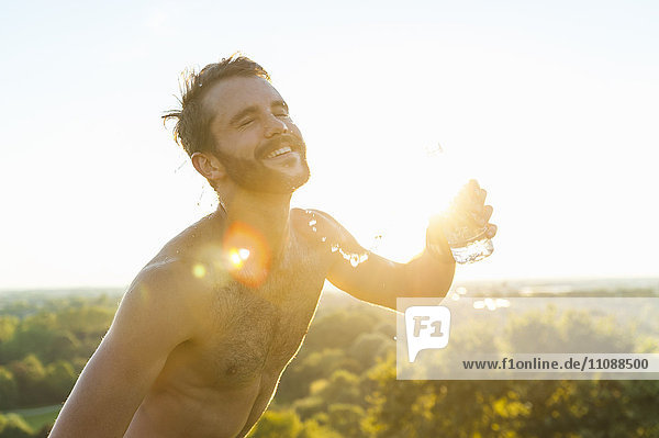 Glücklicher nackter Athlet  der bei Sonnenuntergang Wasser über seinen Körper gießt.