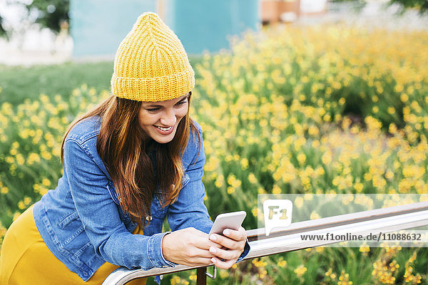 Lächelnde junge Frau mit gelber Mütze beim Blick aufs Handy