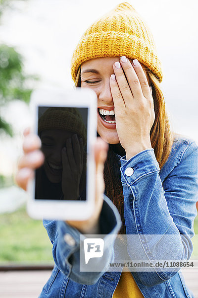 Porträt einer lachenden jungen Frau  die ein Smartphone mit eigenem Bild zeigt