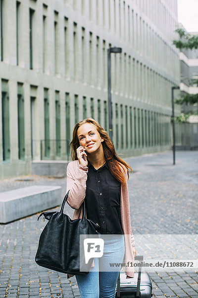 Smiling young woman with wheeled luggage and leather bag on the phone