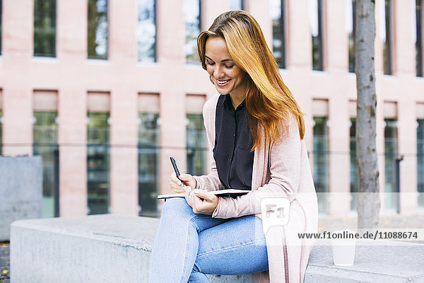 Lächelnde junge Frau sitzt auf Bank mit Notizbuch und Kaffee zum Mitnehmen