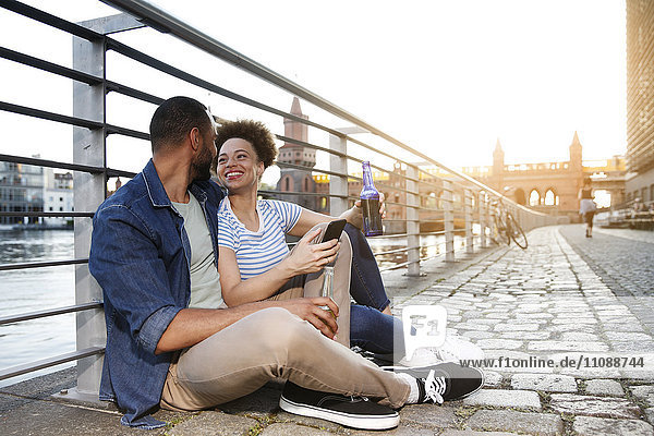 Couple sitting by river holding bottles and smart phone