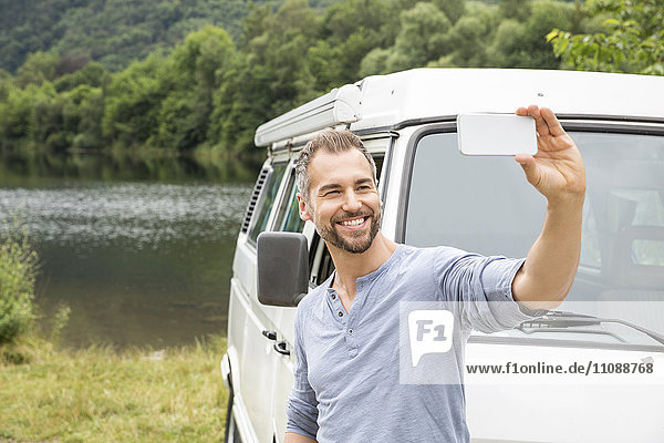Lächelnder Mann mit einem Selfie an einem Van am Seeufer