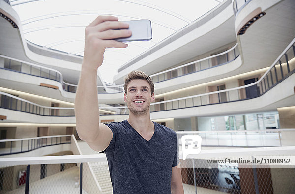Lächelnder junger Mann  der einen Selfie in einem modernen Bürogebäude nimmt.