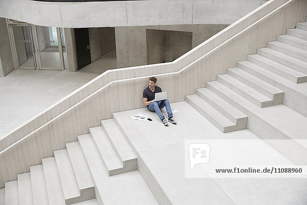 Junger Mann sitzt auf der Treppe im Bürogebäude mit Laptop