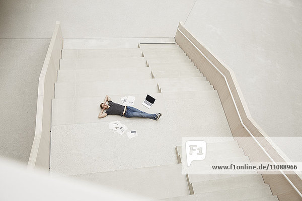Junger Mann auf der Treppe liegend mit Laptop und Papieren