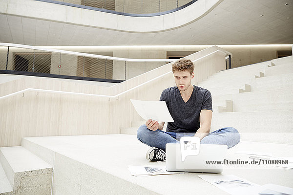 Junger Mann auf dem Boden sitzend mit Laptop und Papieren