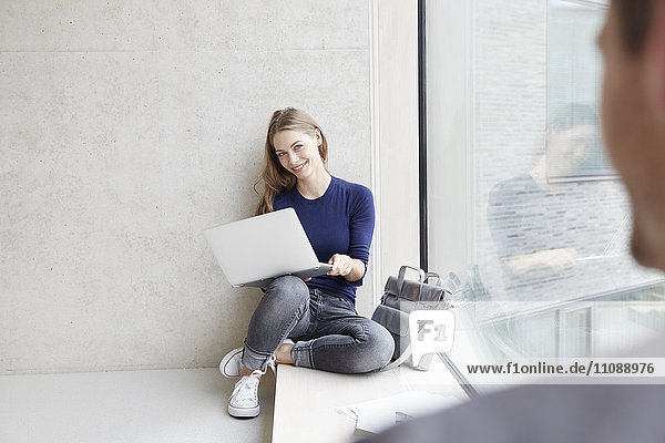 Lächelnde junge Frau an der Betonwand sitzend mit Laptop und Blick auf den Mann