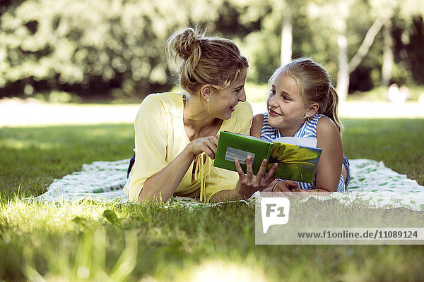 Mädchen und junge Frau mit Buch auf Decke im Park liegend