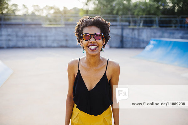 Porträt einer lächelnden jungen Frau in einem Skatepark mit verspiegelter Sonnenbrille