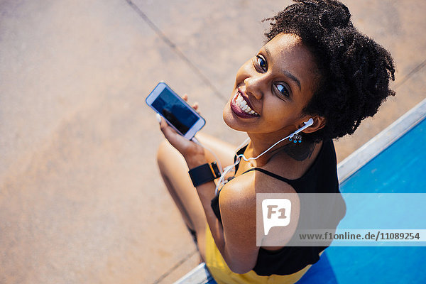 Lächelnde junge Frau mit Kopfhörer und Handy im Skatepark mit Blick auf die Kamera