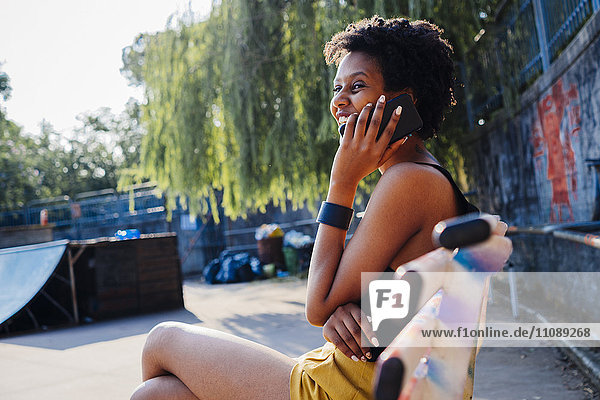 Glückliche junge Frau sitzt auf einer Bank im Skatepark und telefoniert mit dem Handy.