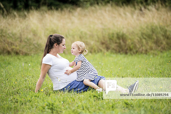 Pregnant woman sitting in grass with little daughter touching mother's belly