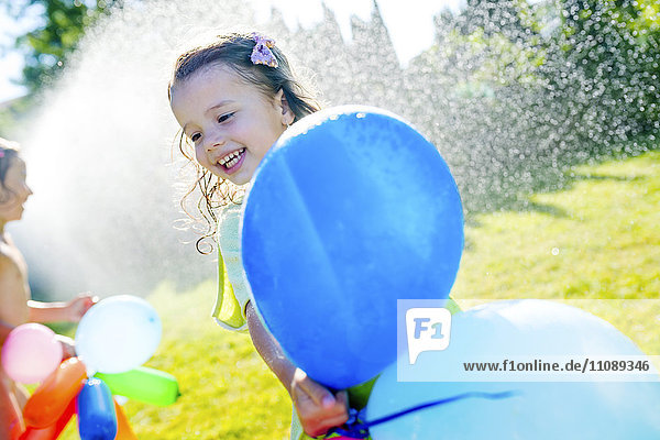 Little girl with balloons having fun with lawn sprinkler in the garden