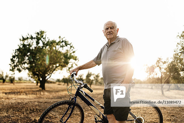 Senior man with bicycle standing on a field at evening twilight