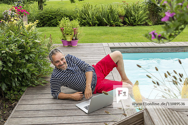 Smiling man sitting at pool edge with his laptop