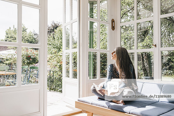Frau sitzend mit Buch zur Lounge im Wintergarten mit Blick durch geöffnete Terrassentür