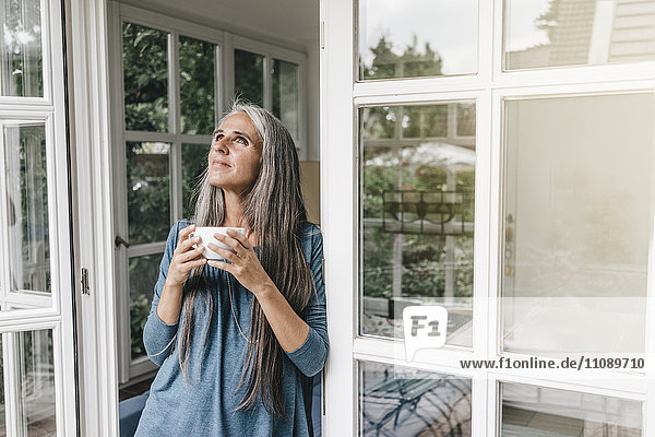 Lächelnde Frau  die sich an den Türrahmen ihres Wintergartens lehnt und mit einer Tasse Kaffee nach oben schaut.