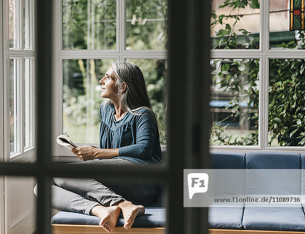 Relaxed woman sitting with book on lounge in winter garden looking through window