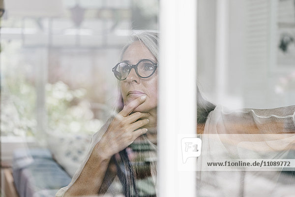 Nachdenkliche Frau sitzt im Wintergarten und schaut durchs Fenster.