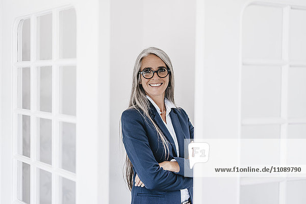 Porträt einer lächelnden Geschäftsfrau mit langen grauen Haaren und Brille zwischen zwei Glastüren.