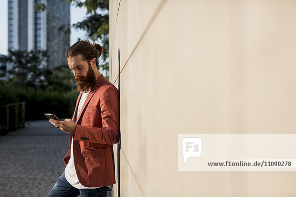 Fashionable young man leaning against wall looking at smartphone