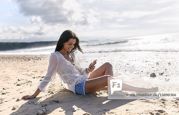 Spain  Asturias  beautiful young woman with smartphone on the beach