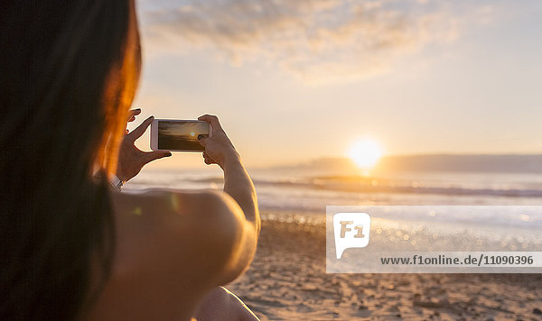 Spanien  Asturien  schöne junge Frau beim Fotografieren am Strand bei Sonnenuntergang  Rückansicht