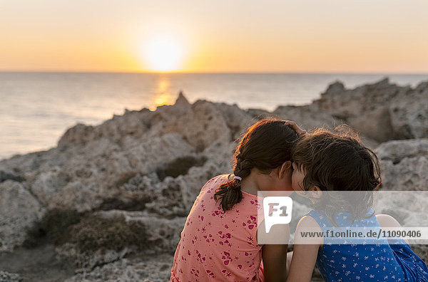 Back view of two little sisters sitting at rocky coast by sunset