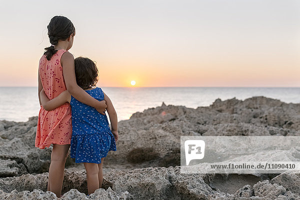 Rückansicht von zwei kleinen Schwestern  die Arm in Arm an der felsigen Küste stehen und den Sonnenuntergang beobachten.