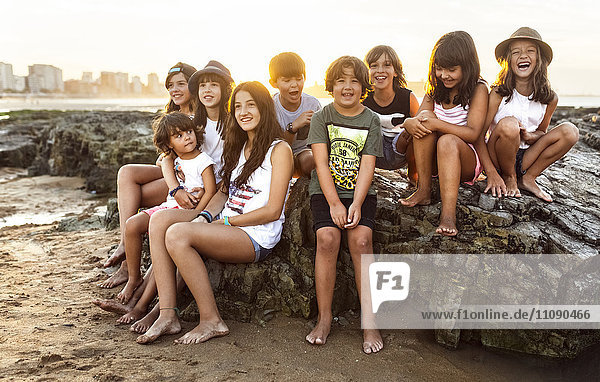 Gruppe von Kindern  die bei Sonnenuntergang auf einem Felsen am Strand sitzen.