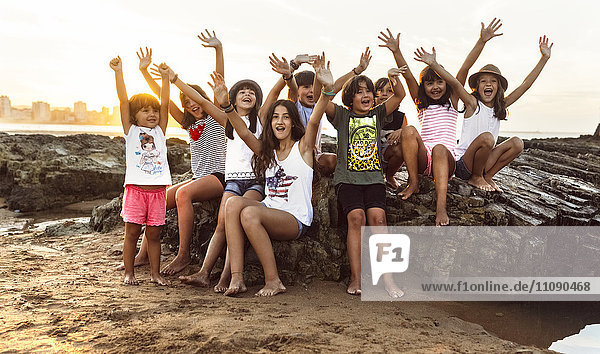 Gruppe von fröhlichen Kindern  die bei Sonnenuntergang auf einem Felsen am Strand sitzen