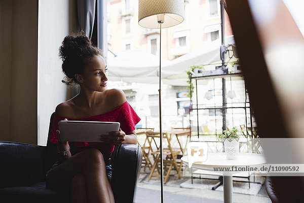 Nachdenkliche junge Frau mit Tablette  die in einem Café sitzt und durchs Fenster schaut.