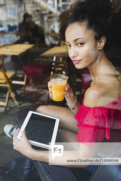 Junge Frau mit einem Glas Orangensaft und Tablette  die durch das Fenster eines Coffeeshops schaut