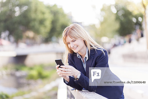 Lächelnde Geschäftsfrau  die sich auf das Geländer stützt und auf das Handy schaut.