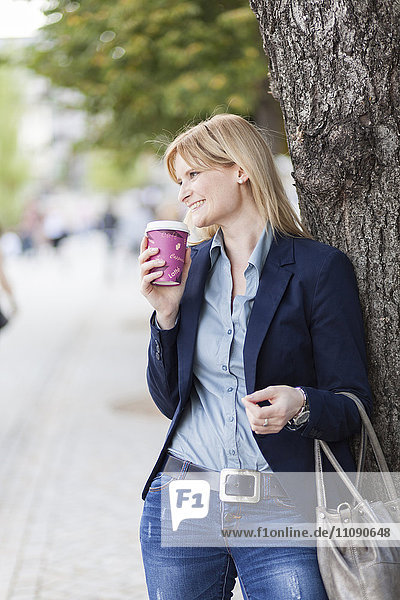 Lächelnde blonde Geschäftsfrau mit Kaffee zum Anlehnen an Baumstamm